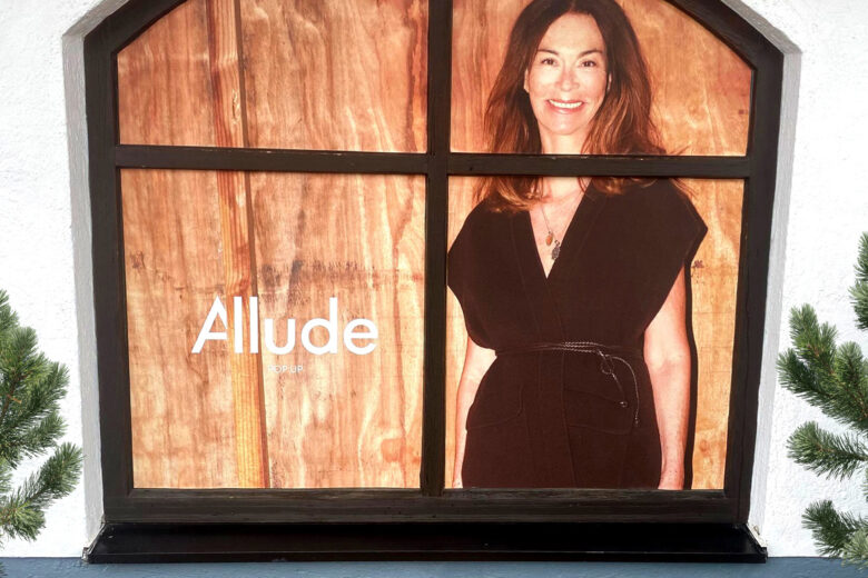 Werbeplakat von Allude mit einem Model in einem schwarzen Kleid vor einer Holzverkleidung.