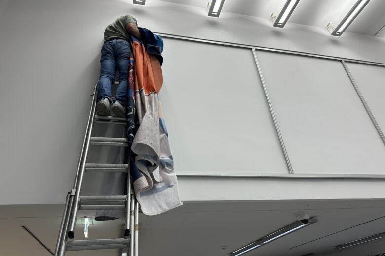 Ein Mitarbeiter montiert einen Textilspannrahmen auf einer Leiter in einem Innenraum.