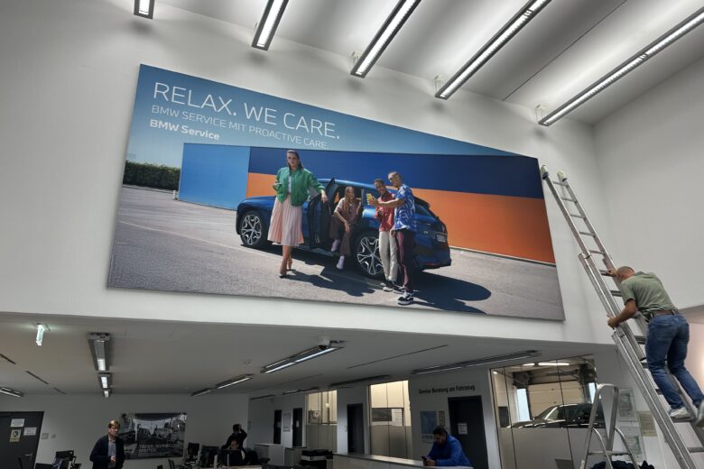 Wandbild im BMW Servicebereich mit dem Slogan 'Relax. We care.' und einer Gruppe von Personen neben einem BMW-Fahrzeug.