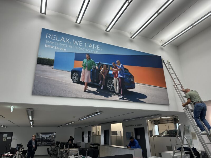 Wandbild im BMW Servicebereich mit dem Slogan 'Relax. We care.' und einer Gruppe von Personen neben einem BMW-Fahrzeug.