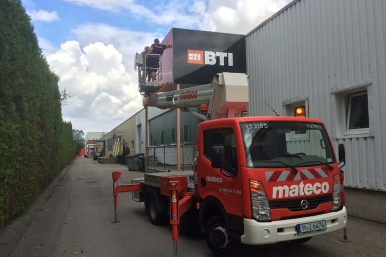 Ein mateco Arbeitsbühne mit einem BTI Firmenschild, das an einem Gebäude angebracht wird.