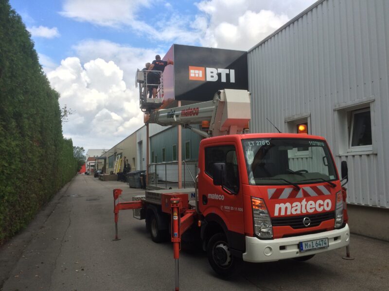 Ein mateco Arbeitsbühne mit einem BTI Firmenschild, das an einem Gebäude angebracht wird.