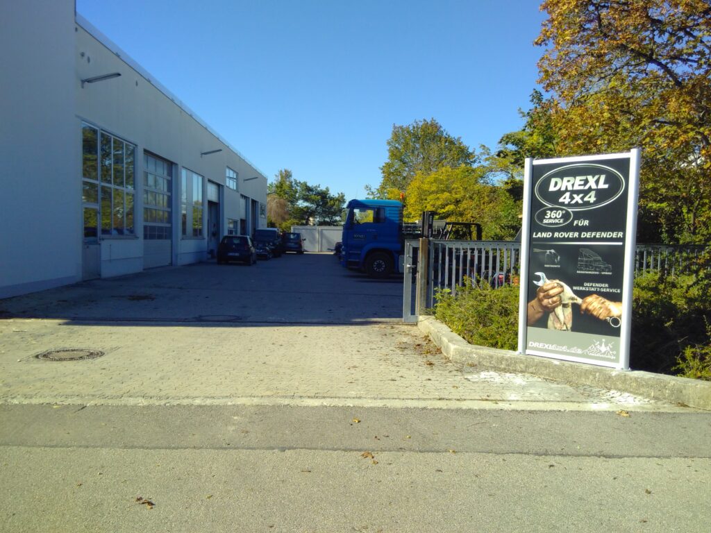 Werbeschild von DREXL 4x4 für den Land Rover Defender an einem Parkplatz.