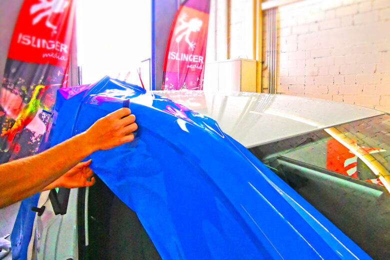 Ein blauer Folier-Rakel wird an einem Fahrzeug angewendet, während ISLINGER-Werbefahnen im Hintergrund sichtbar sind.
