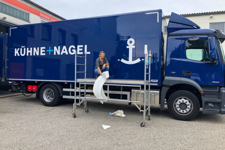 Blauer LKW von KÜHNE + NAGEL mit Folierung und einer Person auf einer Leiter.