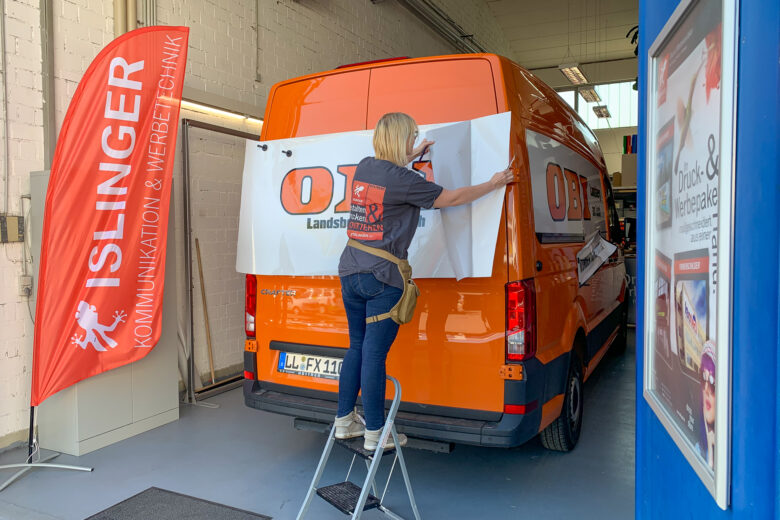 Eine Person bringt eine Fahrzeugbeschriftung mit dem OBI-Logo auf einem orangefarbenen Transporter an.