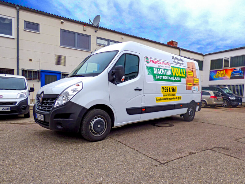Weißer Renault Transporter mit Werbebeschriftung auf der Seite, geparkt auf einem Parkplatz.