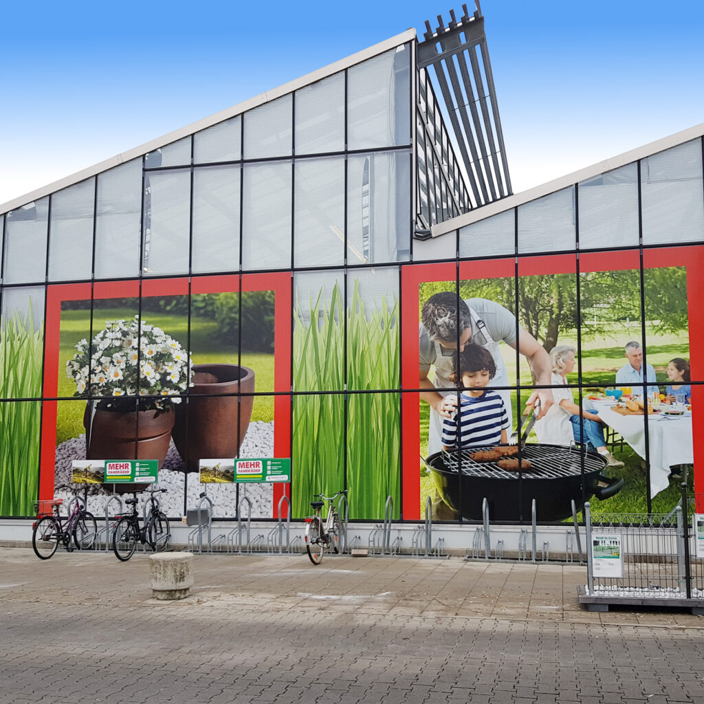 Außenansicht eines Hagebaumarkts mit großen Werbebildern von Pflanzen und Grillaktivitäten.