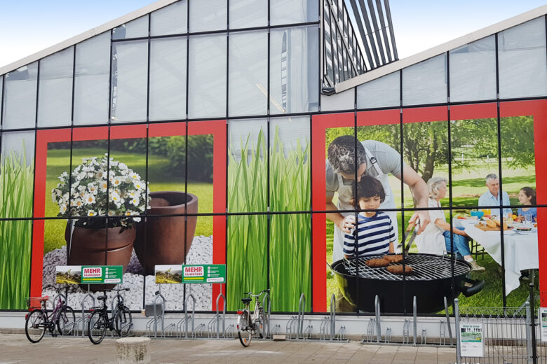Außenansicht eines Hagebaumarkts mit großen Werbebildern von Pflanzen und Grillaktivitäten.