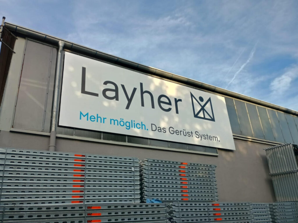 Layher Werbeschild mit dem Slogan 'Mehr möglich. Das Gerüst System.' an einer Wand.