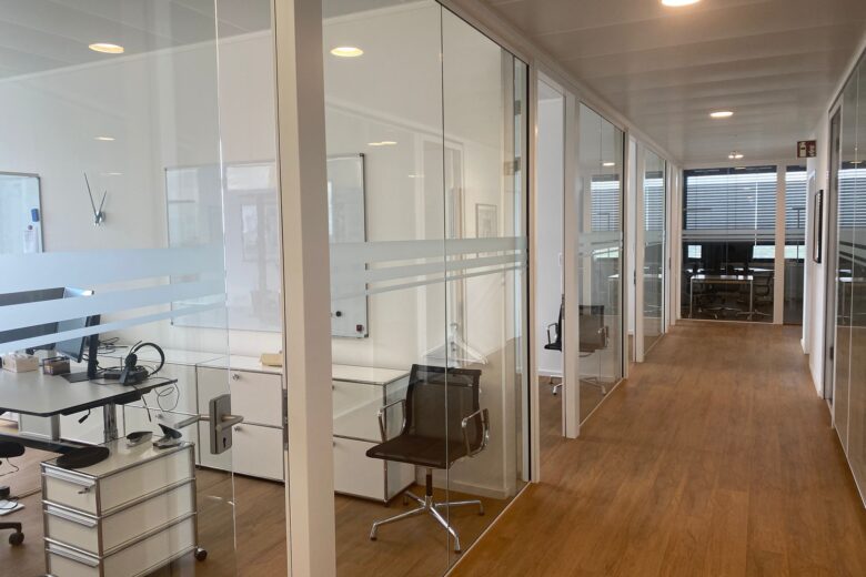 Büroflur mit Glaswänden, Schreibtisch und modernen Bürostühlen in einem hellen Raum.