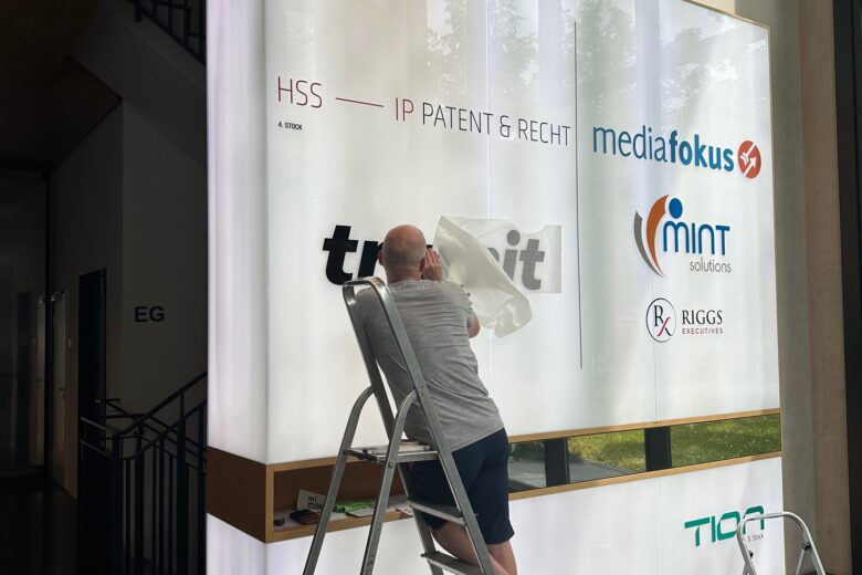 Ein Mann bringt eine Folie auf einen beleuchteten Leuchtkasten mit Unternehmenslogos im Büro an.