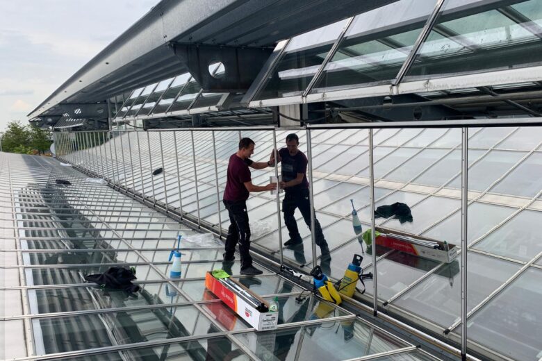 Zwei Arbeiter installieren Sonnenschutzfolierung auf einem Glasdach.