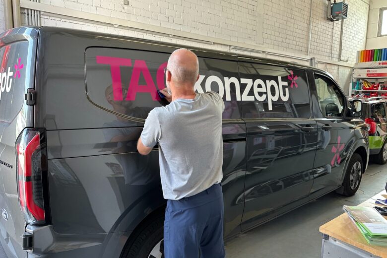 Ein Mann bringt eine Beschriftung an einem grauen Fahrzeug mit dem Logo von TAGKonzept an.