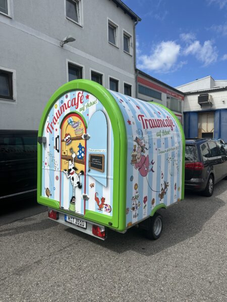 Bunter Verkaufsanhänger des Traumcafes auf Achse mit verschiedenen Illustrationen und einem Fenster für Bestellungen.