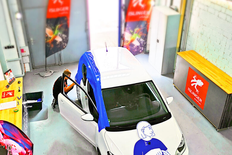 Weißes Auto mit einem blauen Werbebanner und einer Person, die die Tür öffnet, in einem Innenraum.