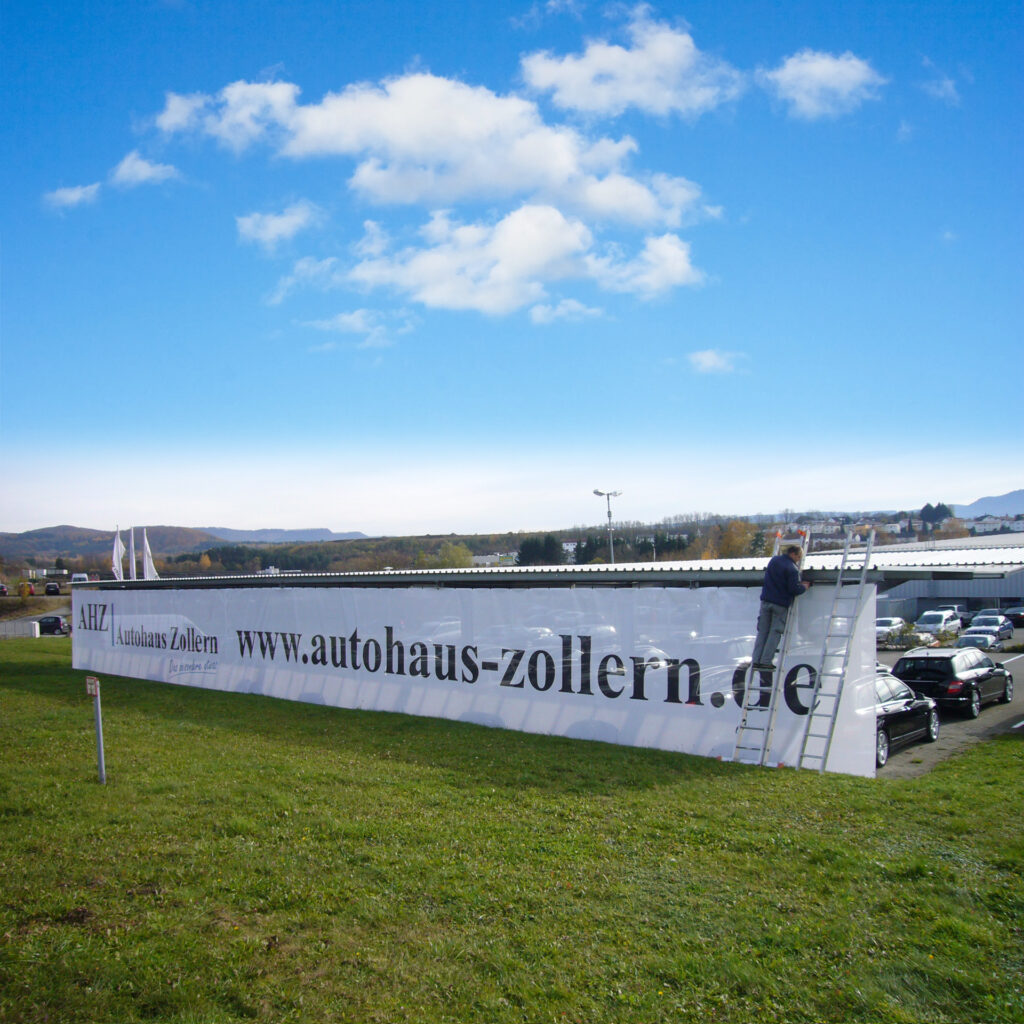 Ein Mitarbeiter montiert ein großes Banner des Autohauses Zollern auf einer Leiter.