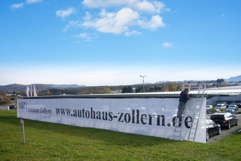 Ein Mitarbeiter montiert ein großes Banner des Autohauses Zollern auf einer Leiter.