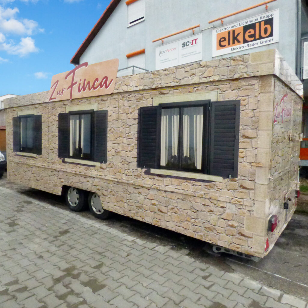 Verkaufsanhänger mit Steinfassade und Fensterläden, beschriftet mit 'Zur Finca'.