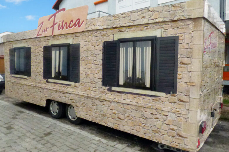 Verkaufsanhänger mit Steinfassade und Fensterläden, beschriftet mit 'Zur Finca'.