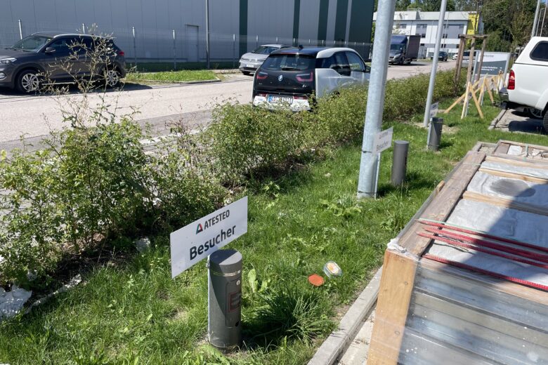 Besucher-Schild von AITESTED auf einem Gehweg neben einer Grünfläche und parkenden Autos.
