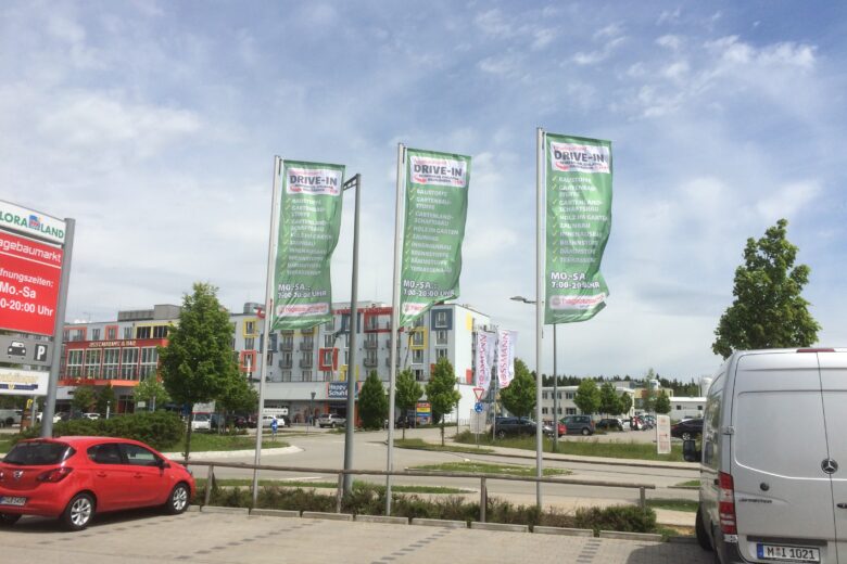 Drei große Werbefahnen für Drive-In, aufgestellt in einem Einkaufsbereich mit Fahrzeugen im Vordergrund.