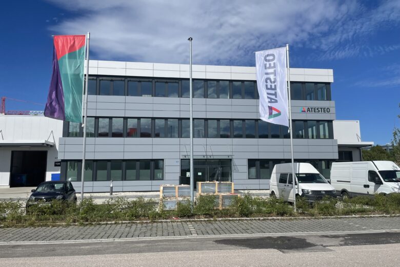 Fassade des ATESTEO Gebäudes mit zwei Fahnen und mehreren Lieferwagen im Vordergrund.