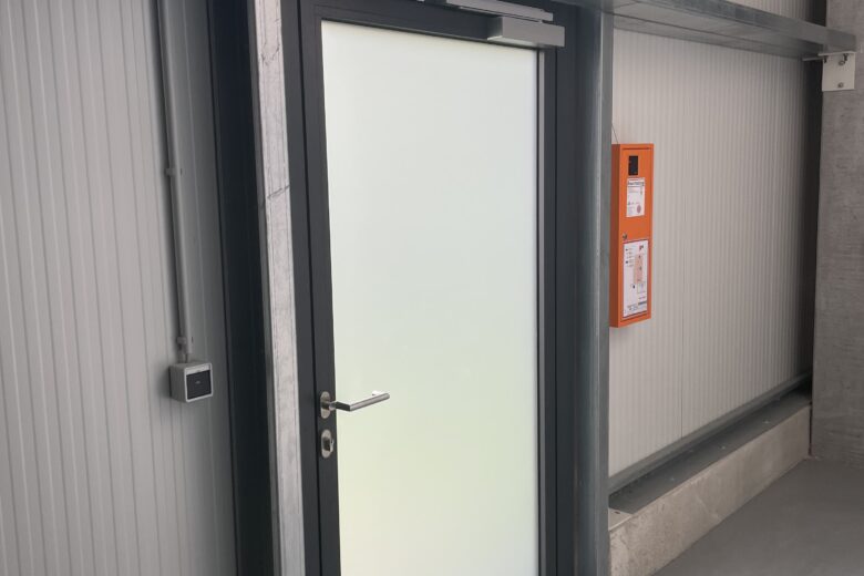 Eine gläserne Tür mit einem Notausgangssymbol und einem Hinweis auf die Notausgangsbeleuchtung.