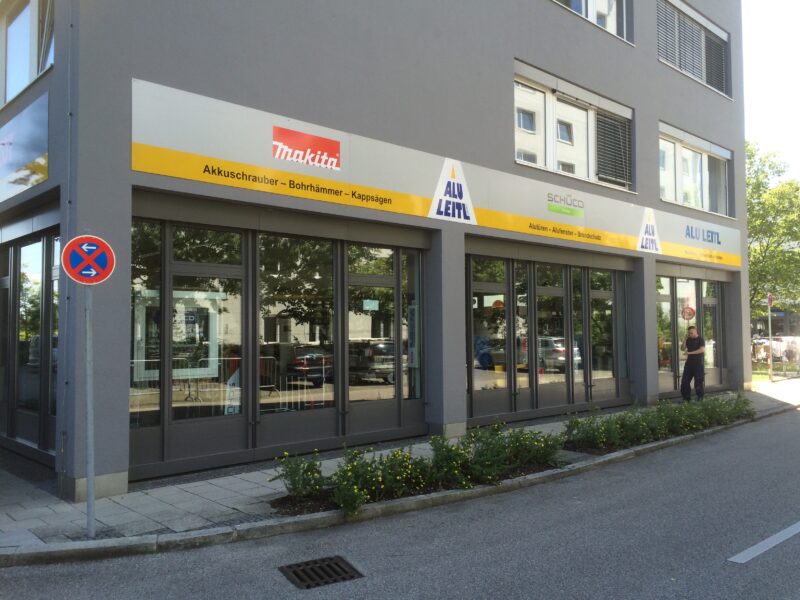 Fassade mit Werbeschildern eines Geschäftes mit gelben Beschilderungen von Makita, SCHÜCO und ALU LEITL, die verschiedene Werkzeuge und Dienstleistungen anzeigen.