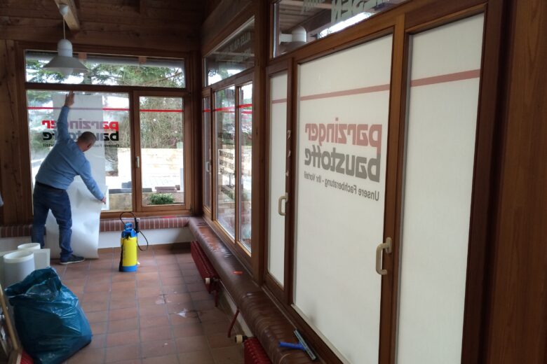 Ein Handwerker bringt eine Fensterfolie an einem großen Fenster an, während er auf einem Tisch steht.