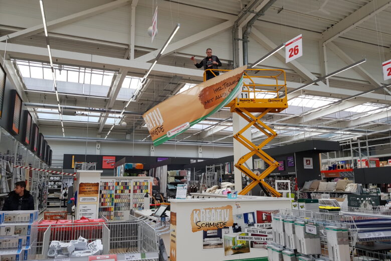 Ein Mitarbeiter auf einer Hebebühne, der ein großes Banner in einem Baumarkt anbringt.