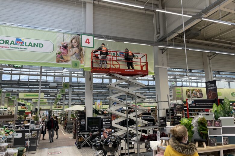 Zwei Mitarbeiter stehen auf einer Hebebühne im Supermarkt und arbeiten an einer oberen Regalfläche.