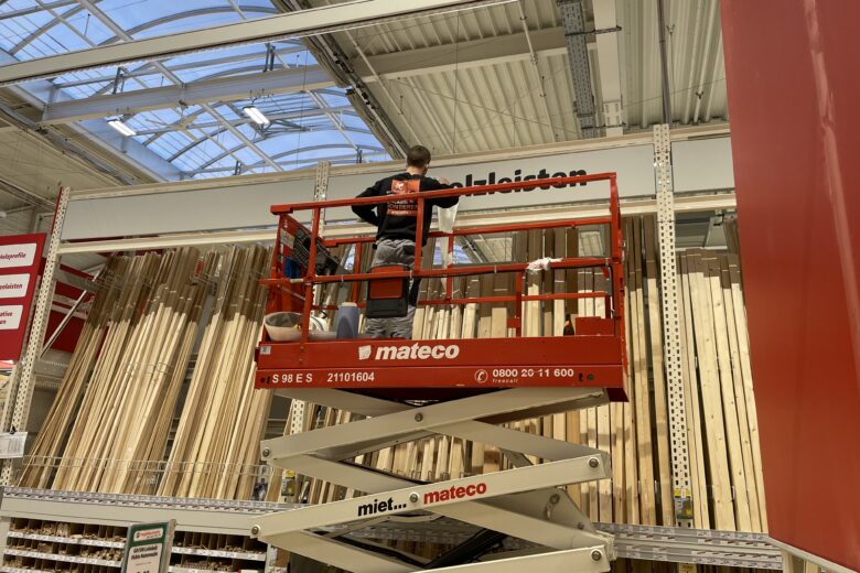 Mitarbeiter steht auf einer Hebebühne und arbeitet an einem Regal im Baumarkt mit Holzlatten.