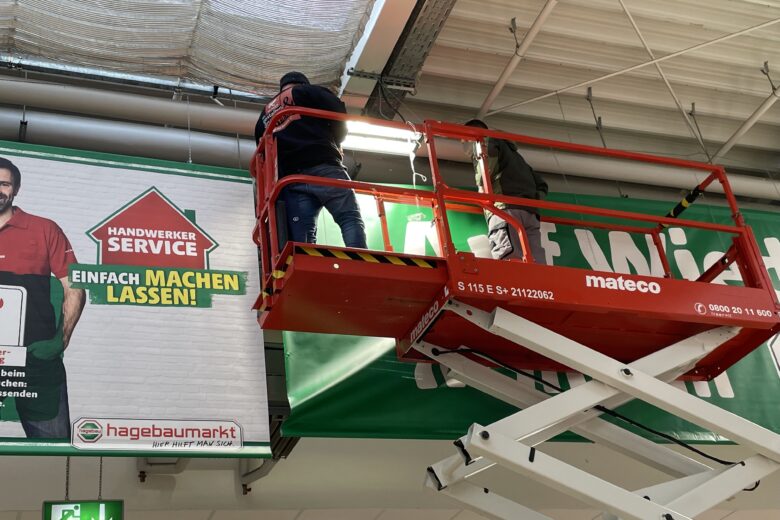 Zwei Mitarbeiter arbeiten mit einer Hebebühne im Inneren eines Baumarkts zur Installation von Beleuchtung.