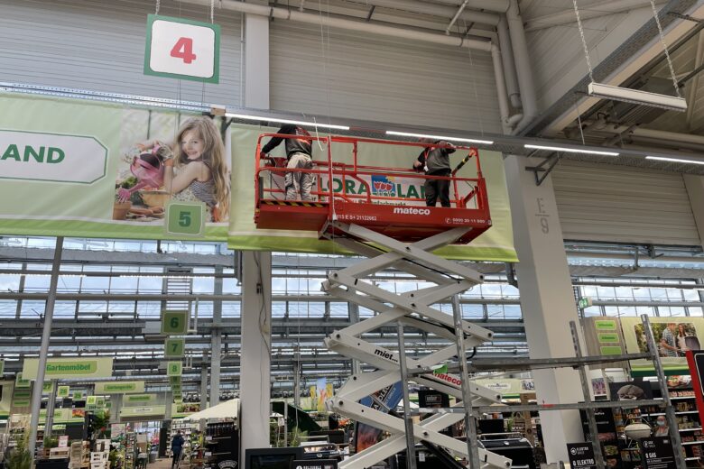 Arbeiter auf einer Hebebühne montieren ein großes Werbebanner in einem Geschäft.
