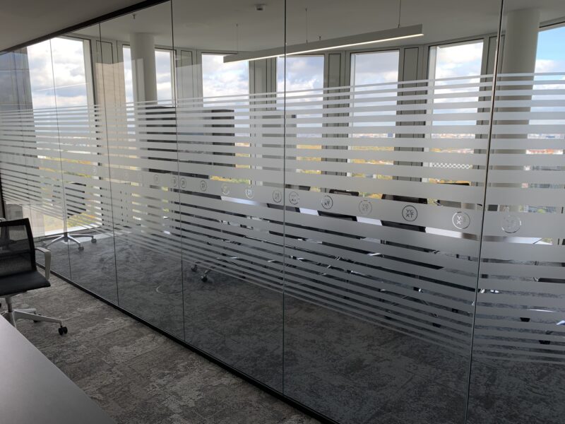 Transparente Glaswand mit horizontalen Streifen und Symbolen in einem modernen Bürogebäude.