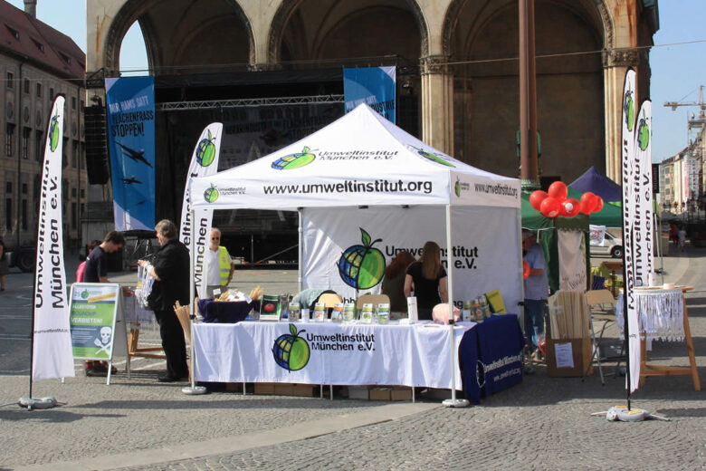 Stand des Umweltinstituts München e.V. mit Tischen und Informationsmaterialien auf einer Veranstaltung.