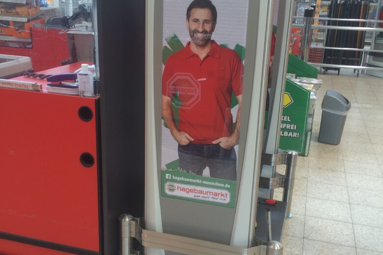Werbesäule mit einem Mann in rotem Hemd und dem Text 'NR. 1 in München' bei einem Checkpoint.