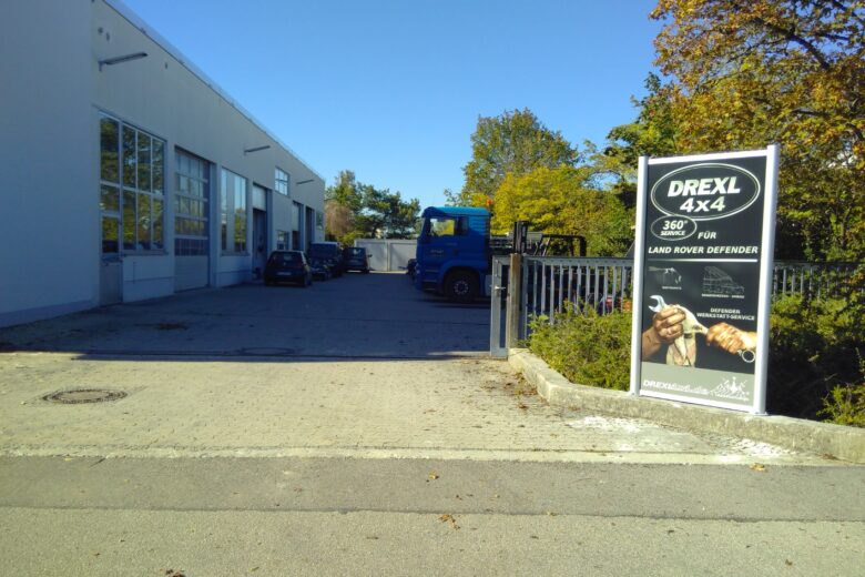 Werbeschild für DREXL 4x4 mit Land Rover Defender und einem Handshake auf einem Parkplatz.