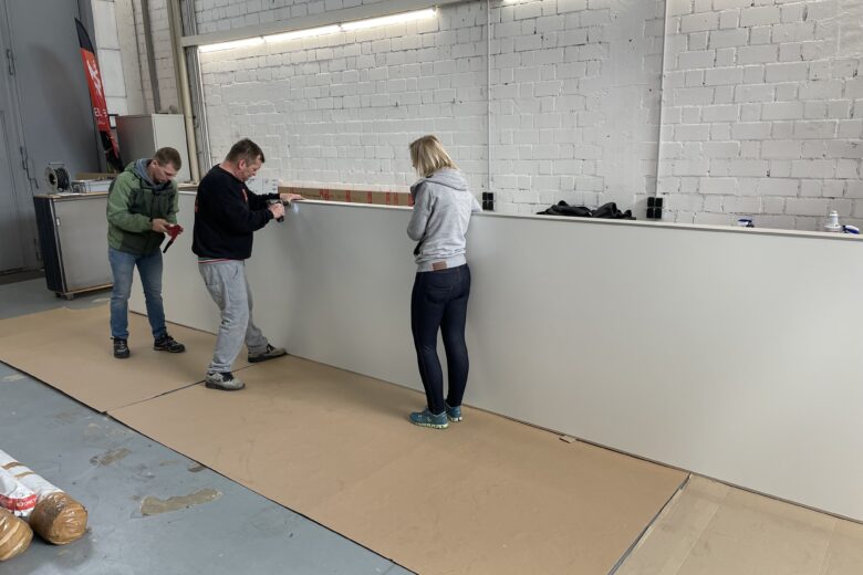 Drei Personen montieren einen Textilspannrahmen in einem Innenraum mit einem neutralen Hintergrund.