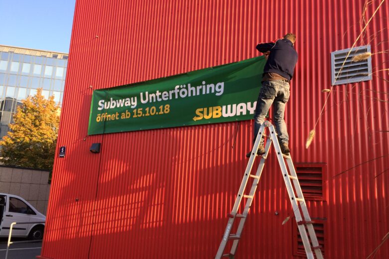 Ein Mann bringt ein Subway-Werbebanner an einer roten Wand an, das das Öffnungsdatum anzeigt.