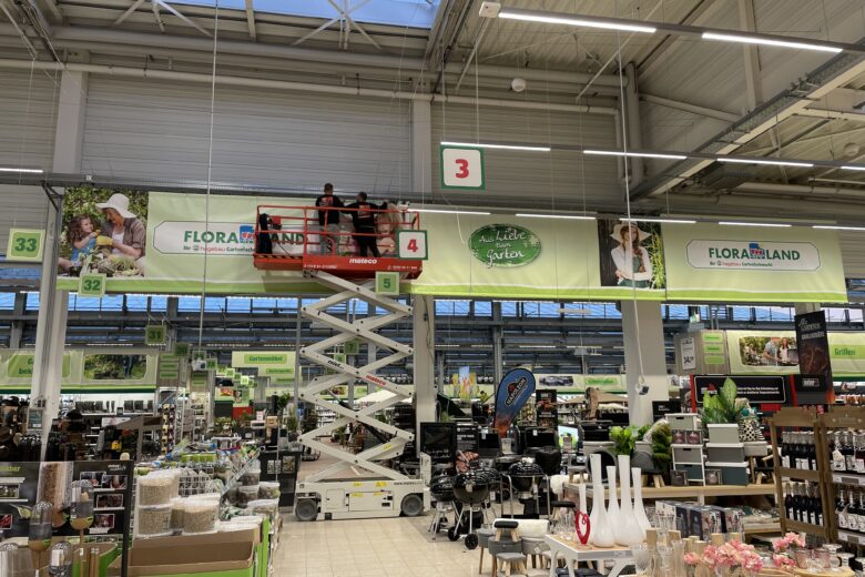 Zwei Personen auf einer Hebebühne, die ein Werbebanner im Floraland Geschäft anbringen.